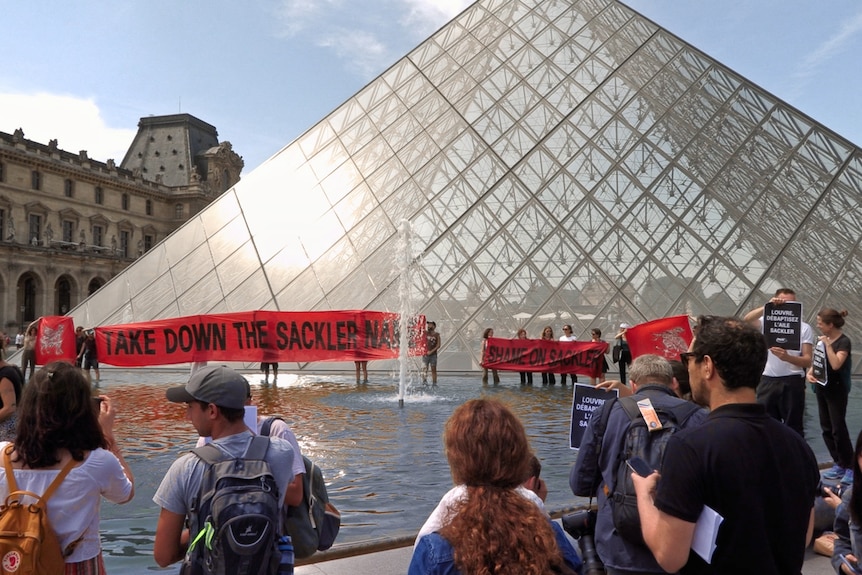 A group of protestors stand holding banners.around the fountain outside the Louvre, a glass pyramid gallery.