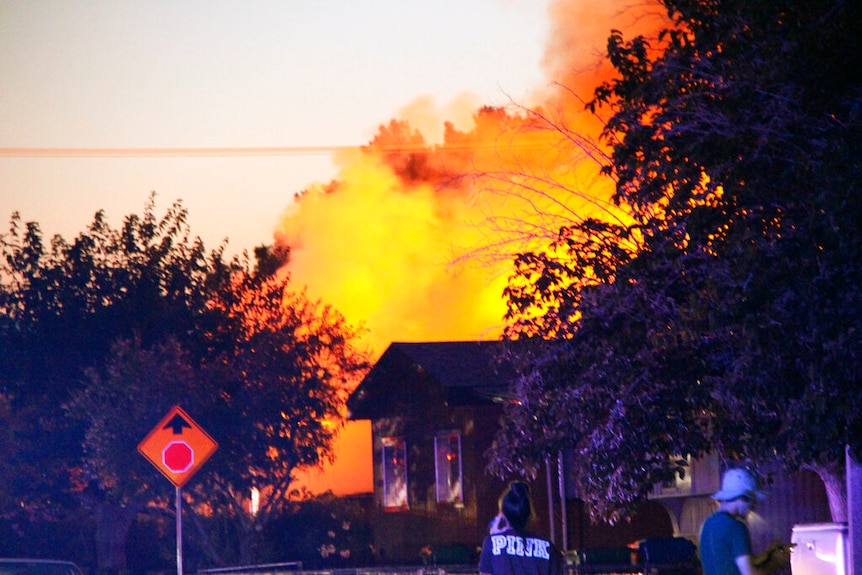 Emergency services attend a large fire that is raging behind a house on a suburban street.