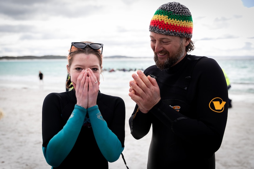 A woman and a man in wetsuits, man wears colourful knitted beanie, woman covers mouth and nose with hands, on beach.