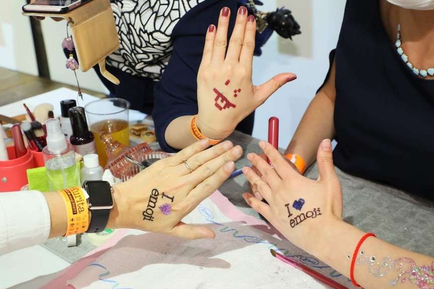 Three women hold up their hands decorated with "I love emoji" writing and glitter.