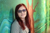 a woman with long red hair and glasses looks toward the camera a she stands with her back to a painted wall