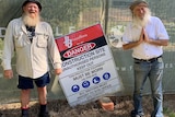 Two older men with beards smile as they stand in front of a fence marked 'Danger: Construction site … keep out'.