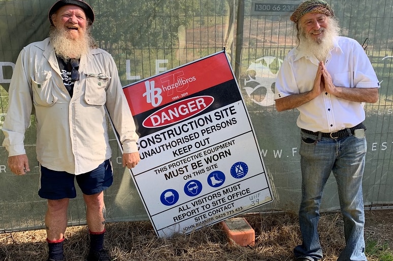 Two older men with beards smile as they stand in front of a fence marked 'Danger: Construction site … keep out'.