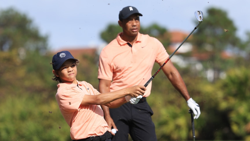 A young golfer plays a tee shot as his dad looks on. They are both dressed the same.