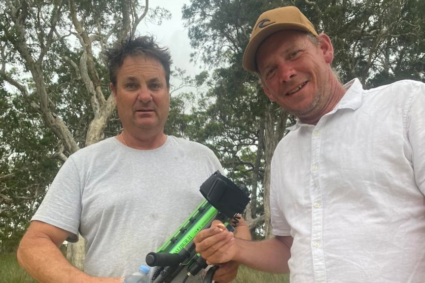 Un homme tenant un détecteur de métaux se tient dans l'eau peu profonde d'une rivière à côté d'un homme tenant une alliance en or.