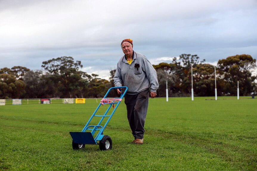 Older man in grey jumper pushes trolley across country footy ground