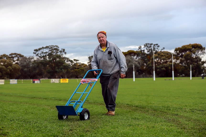 Older man in grey jumper pushes trolley across country footy ground