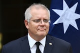 Scott Morrison wearing a black suit and tie in front of an australian flag and a sandstone wall