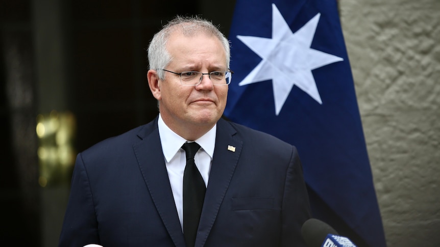 Scott Morrison wearing a black suit and tie in front of an australian flag and a sandstone wall