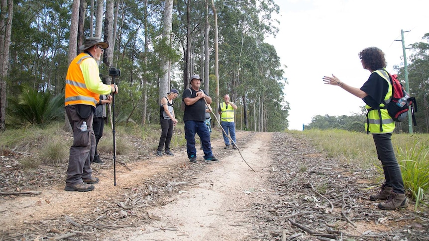 A woman holding gardening gloves and a stick pauses to think on a bush track