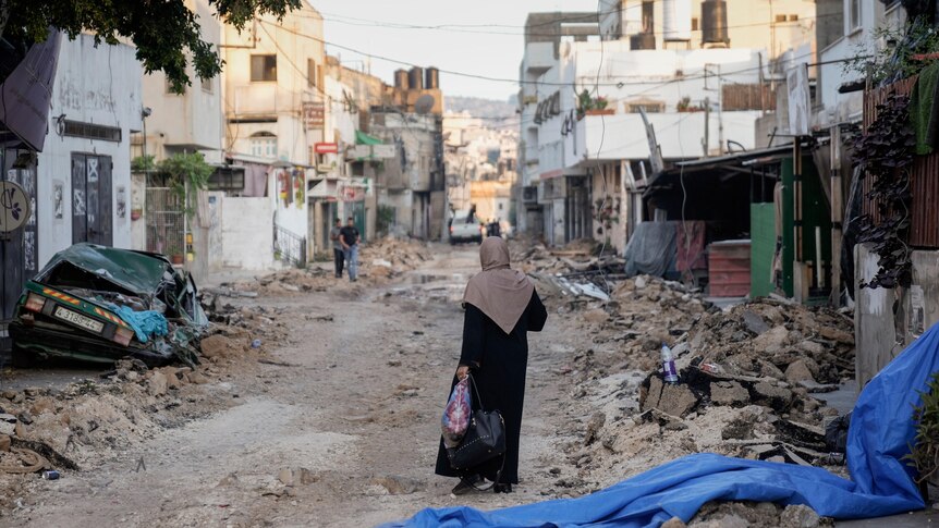 A woman walks on a road with rubble and damaged buildings on both sides of it.