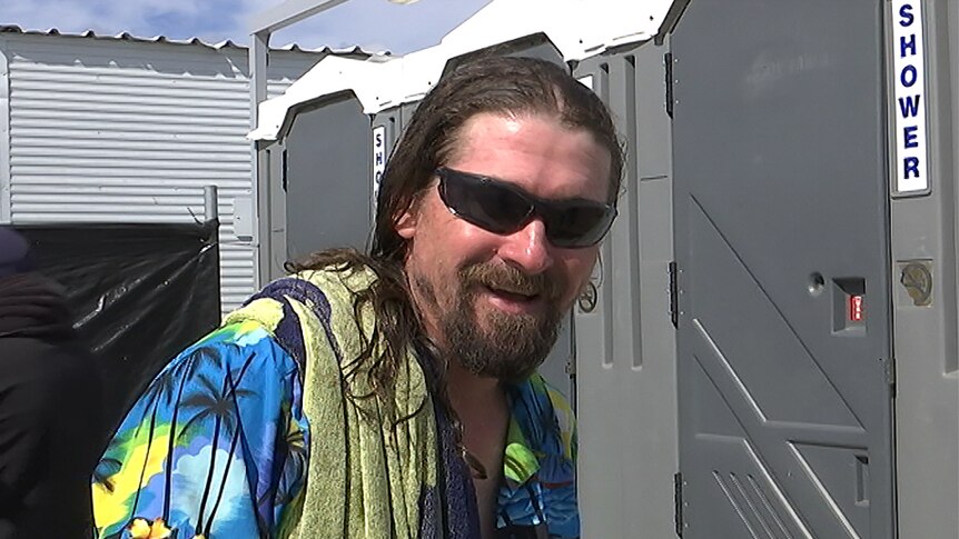 A man with long, wet hair and sunglasses and towel standing in front of shower stalls.