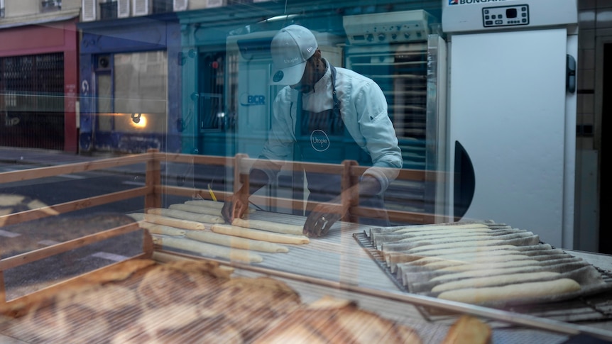 A baker is photographed through a bakery window as he bakes baguettes