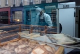 A baker is photographed through a bakery window as he bakes baguettes