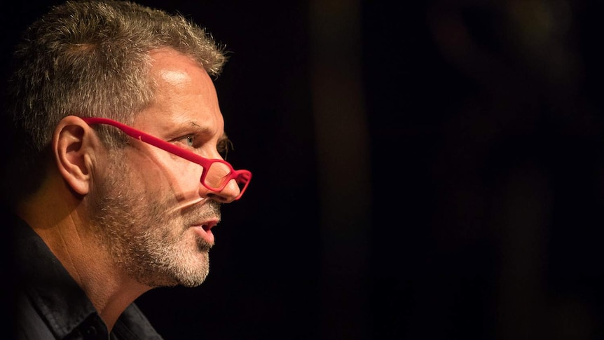 A man, wearing red-rimmed reading glasses, speaks into a microphone