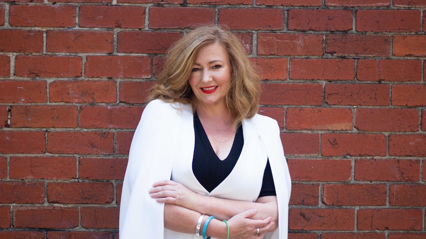 A lady with blonde fuzzy hair and a white shawl looks at the camera while standing against a brick wall.