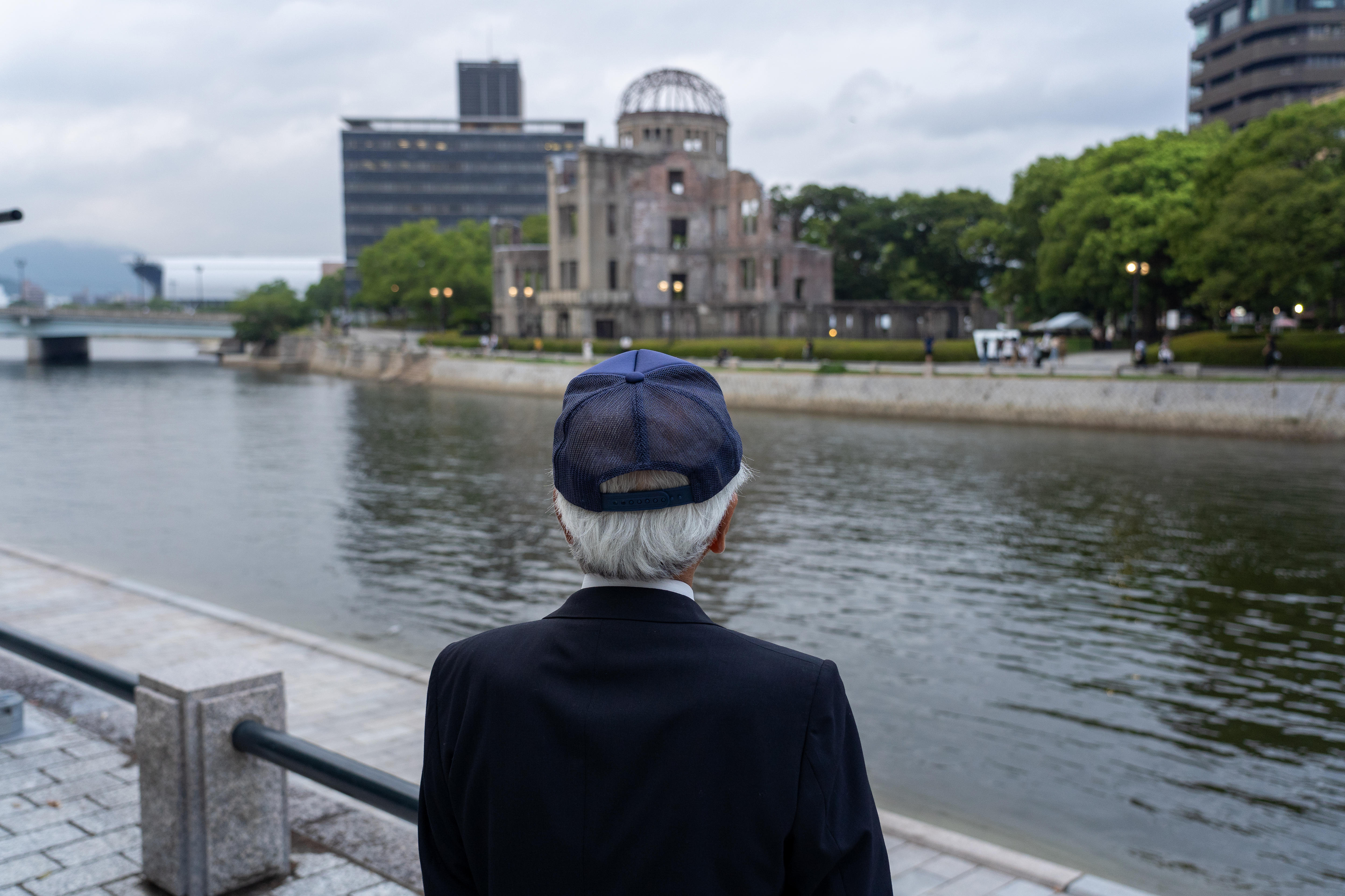 A man with grey hair wearing a dark suit and blue facing away over water, looking at the Hiroshima Peace Dome