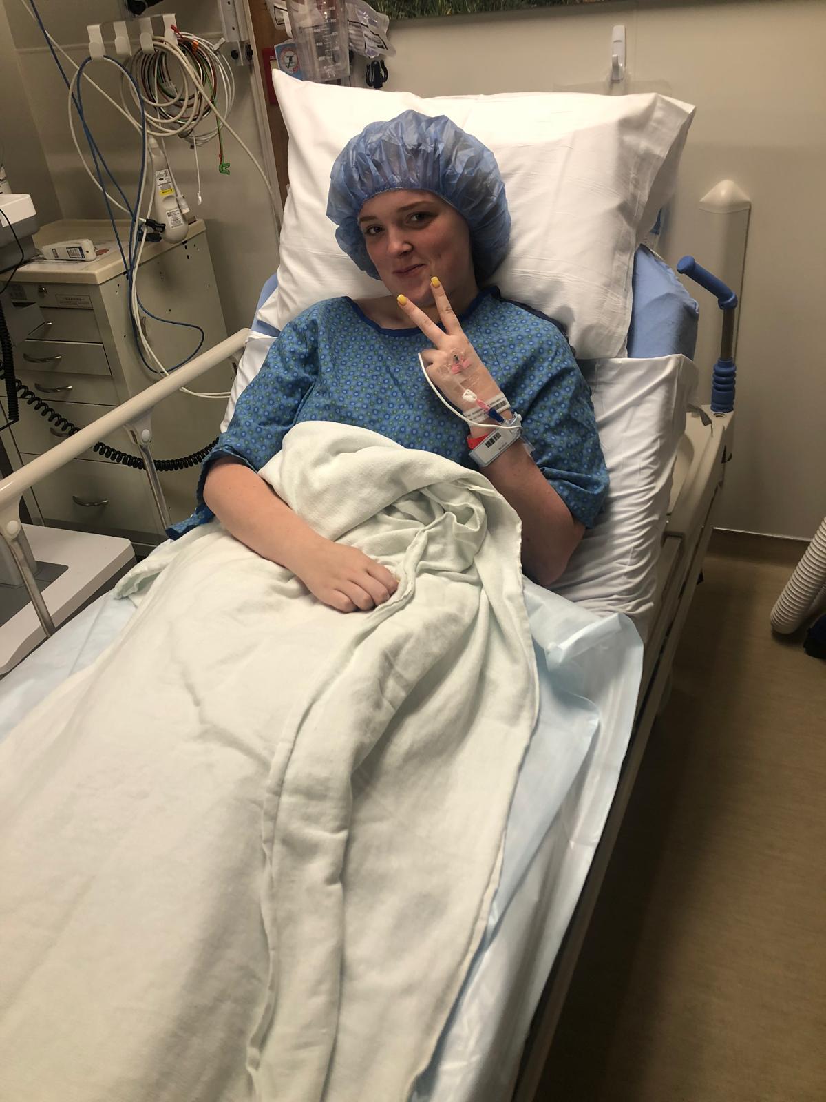 Una joven en pijama azul yace en la cama del hospital, sonriendo y haciendo un signo de paz