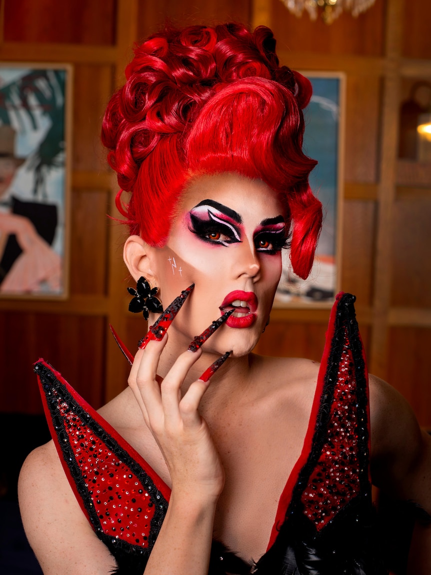 A drag queen wearing a fire-engine red updo wig brings her long red bejewelled nails to her crimson pout