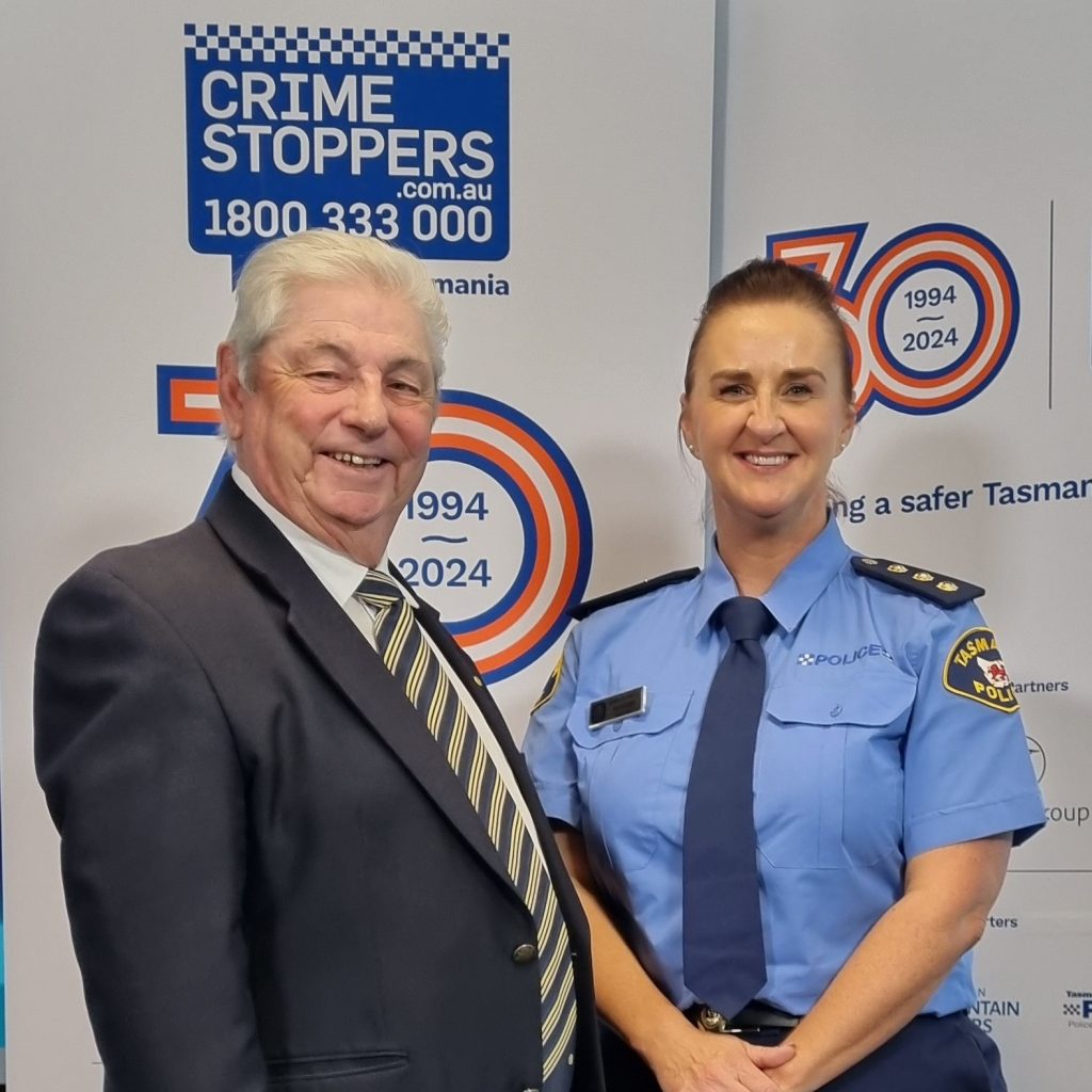 a grey-haired man with a policewoman in front of a crimestoppers logo backdrop