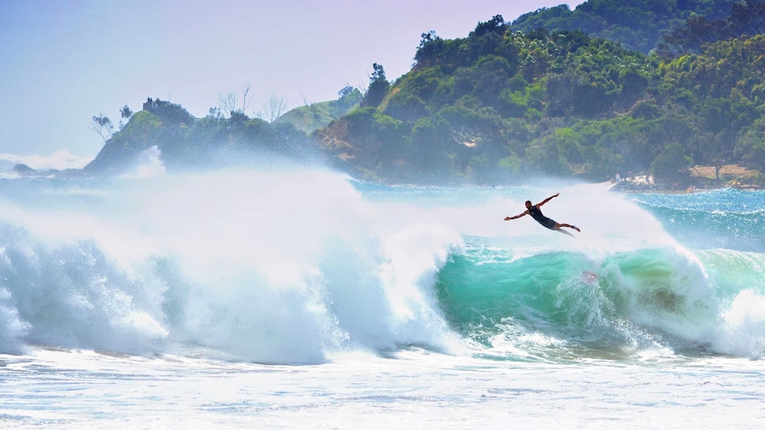 Wide photo of a large wave with a silhouetted surfer flying through the air as he wipes out.