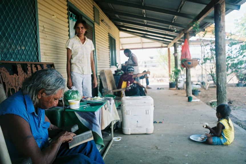 An older woman sits on a porch reading with the rest of her family in the background