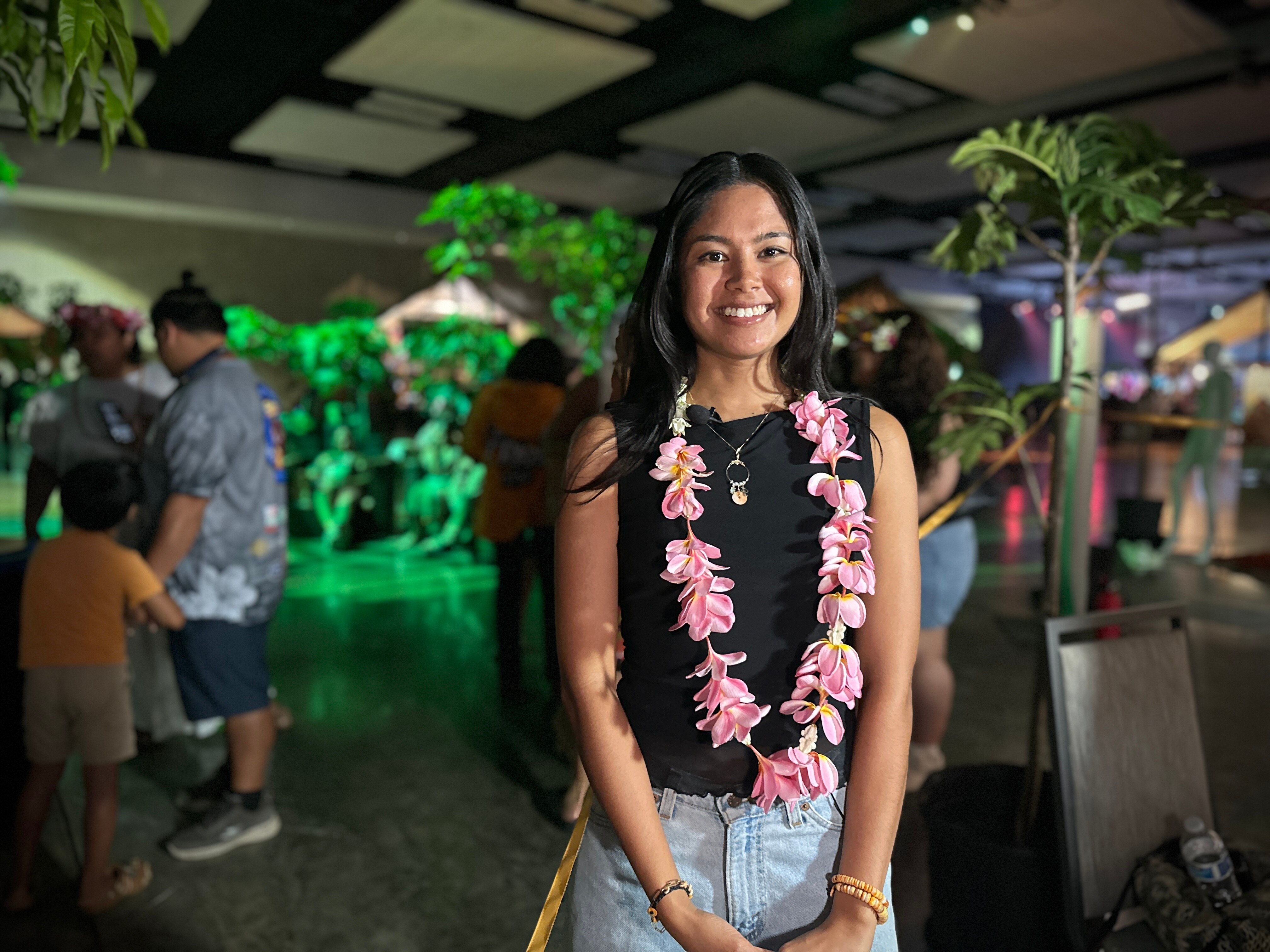 Woman with long black hair stands in busy room. Wears pink flower ula around her neck, black top. 