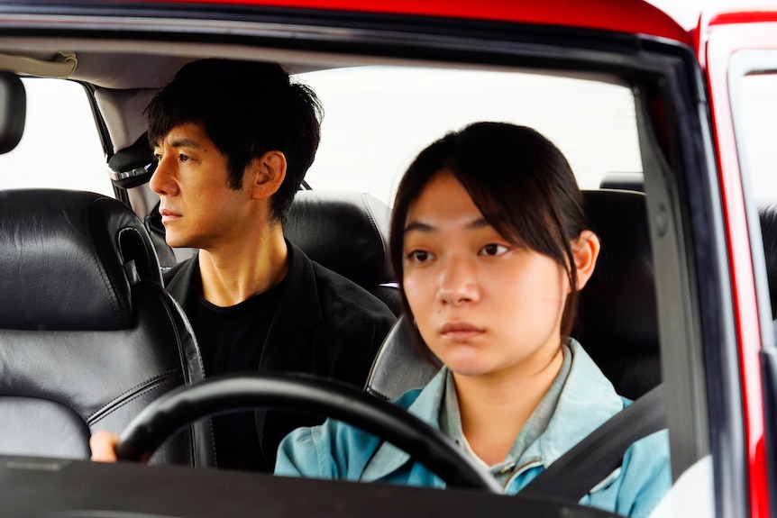 Japanese man with short hair and black shirt sits pensively in back of car while younger Japanese woman in blue shirt drives