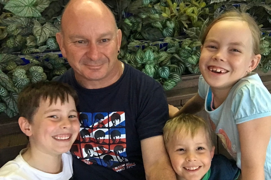 A bald middle-aged man in black T-shirt is surrounded by three smiling young children on wooden bench