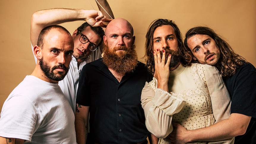 Five members of IDLES stand in front of a brown wall staring at the camera