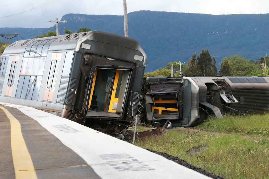 A train carriage lies on its side next to another upright carriage. 