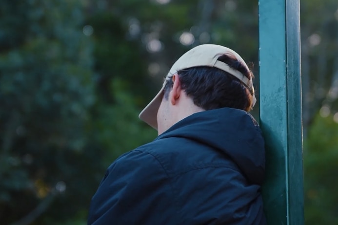 Young man stands facing away from camera wearing a hat 