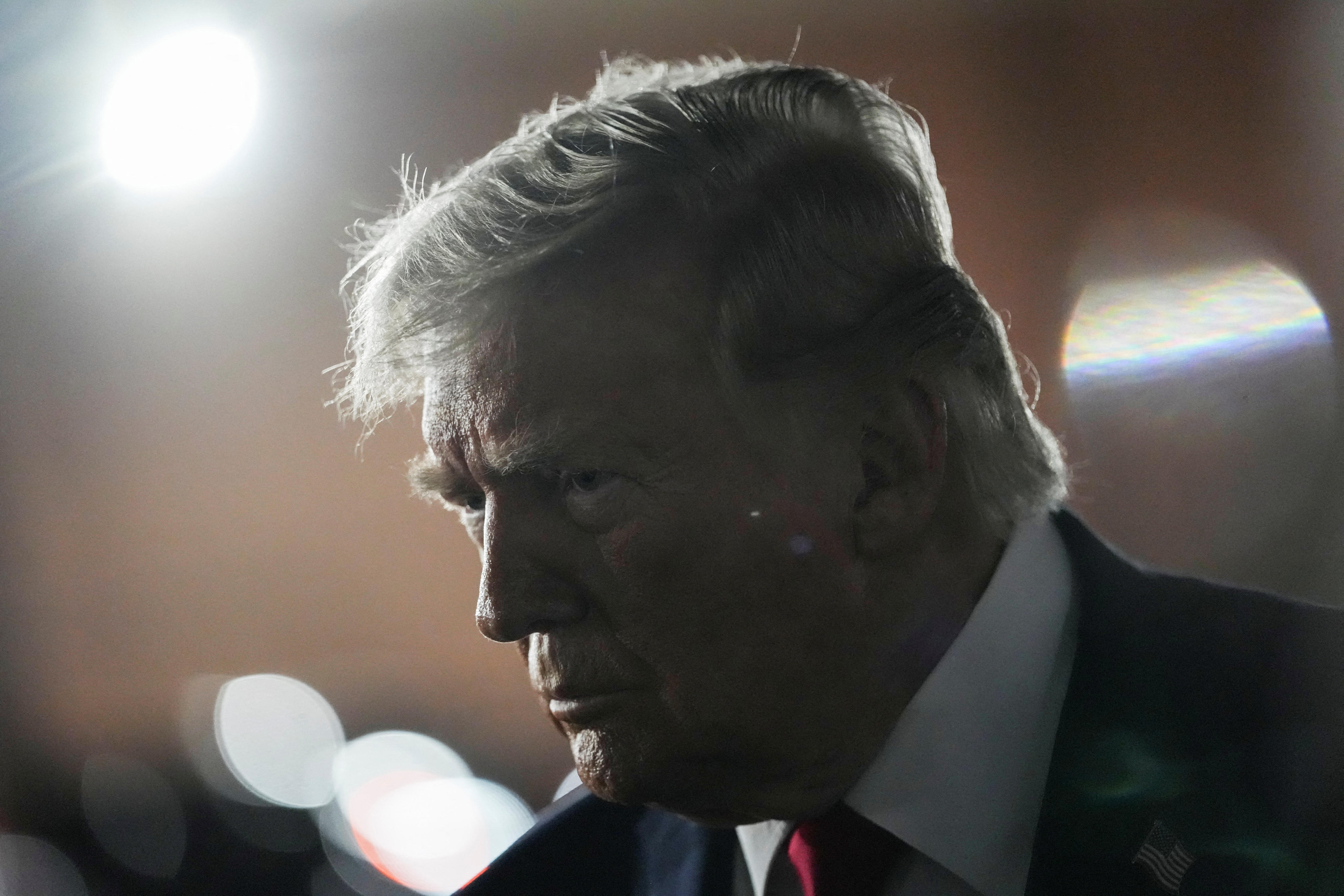 Donald Trump, wearing a suit talks, leans forward. Behind him are bright lights.