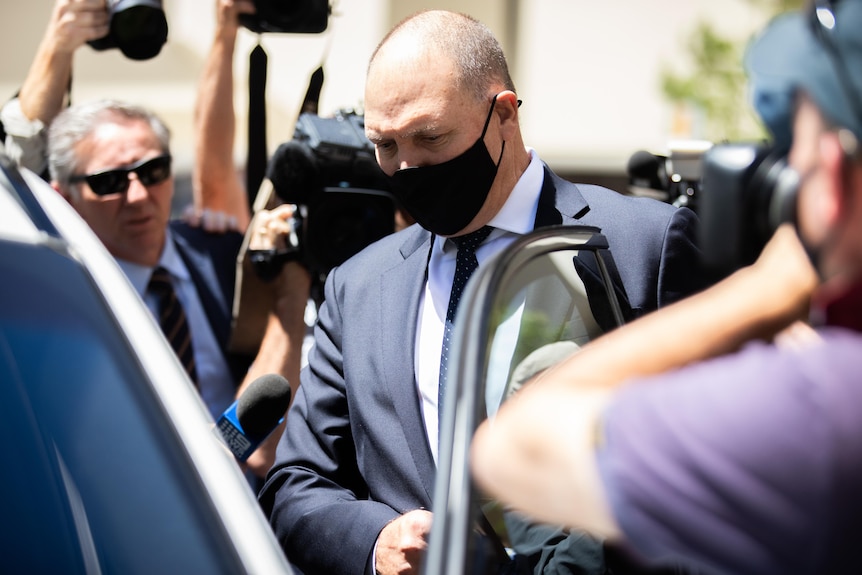 Man wearing a suit and facemask steps into a car, while surrounded by cameramen and journalists.