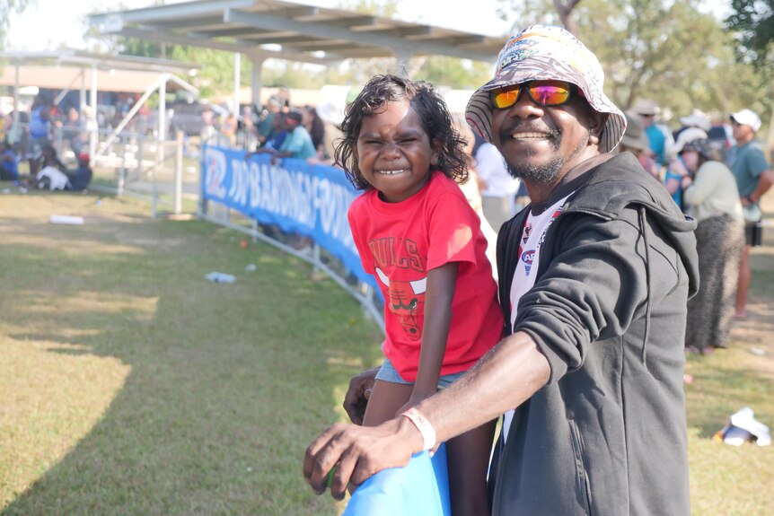 A man and small girl, aged about four, standing behind the barrier of a small football oval.