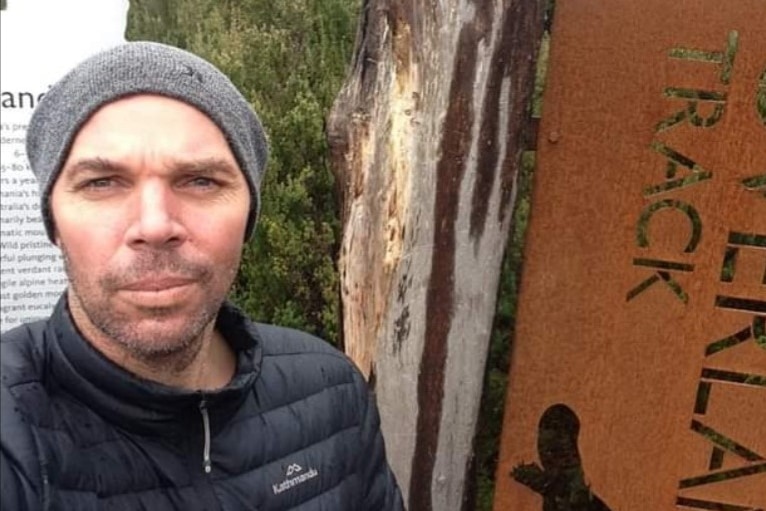 Luke is a wearing a grey beanie standing in front of a tree and sign for a walking track