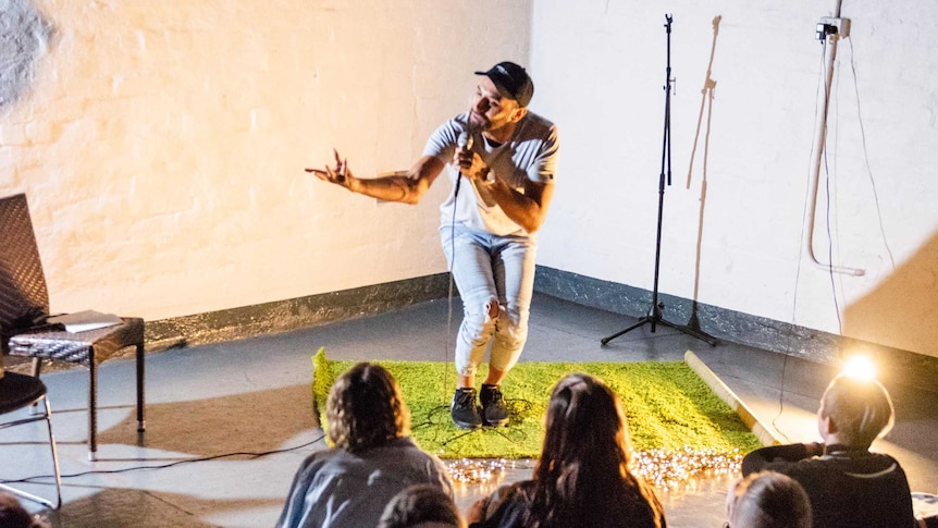 A man holds a microphone and performs in front of students, standing on a small green carpet. Performing as he travels.