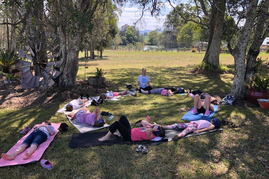 A group of girls lie on mats in a circle on a grassy paddock under some trees.