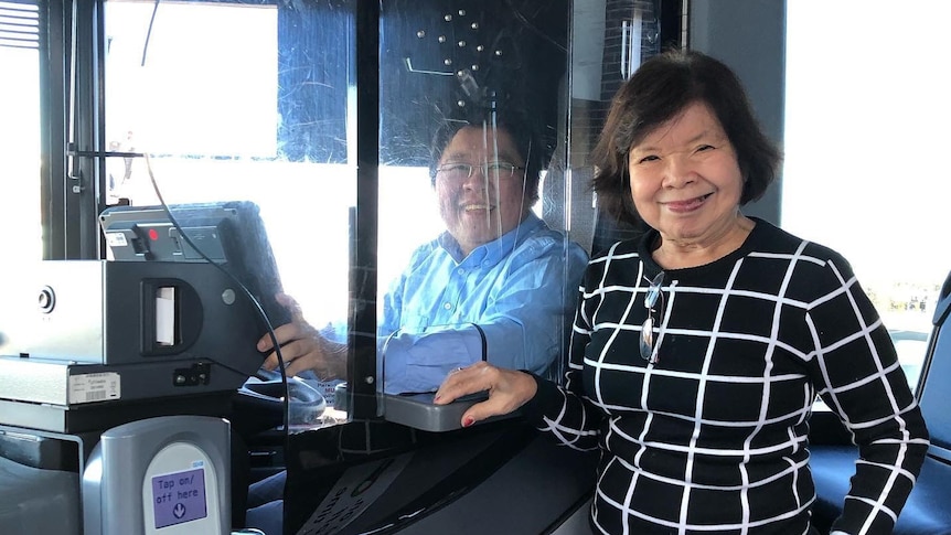 A man sits in a bus driver seat. A woman stands next to him. Both are smiling at the camera.