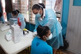 Woman in scrubs and face mask bends to side of young woman sitting on chair.