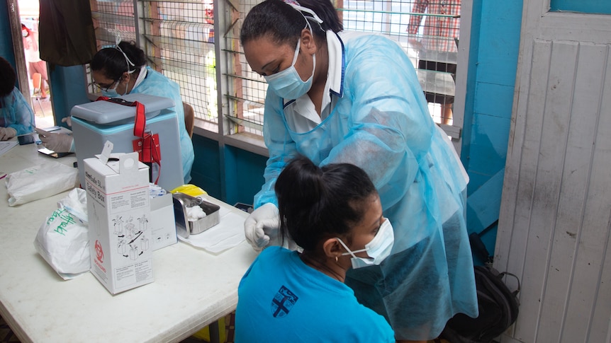 Woman in scrubs and face mask bends to side of young woman sitting on chair.