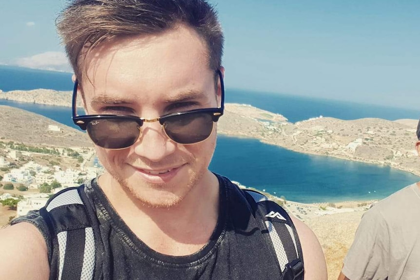 A young man in a black singlet and sunglasses takes a selfie in front of a beach.