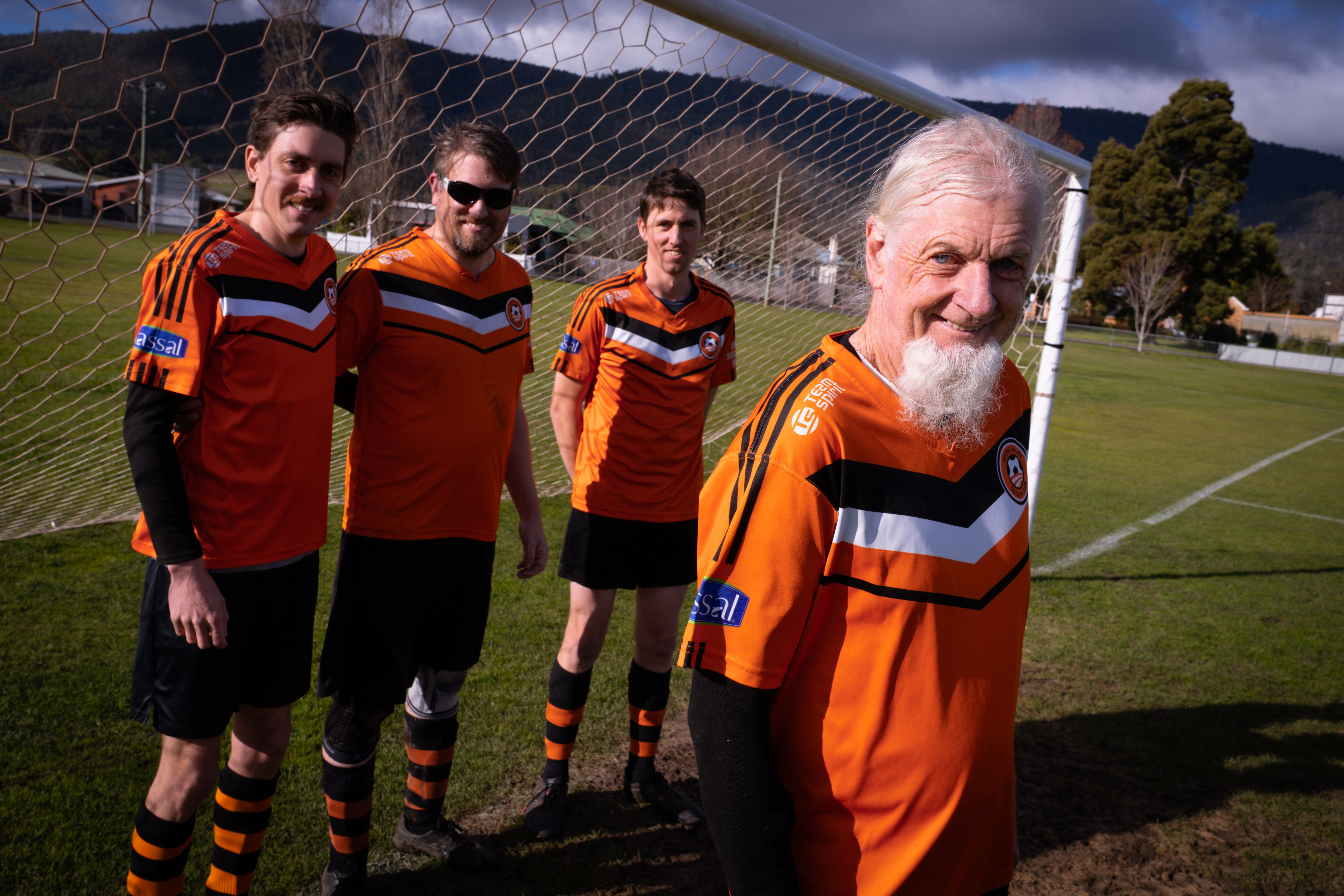 A man with white hair and a long white beard wearing orange soccer colours with three other men in the nets.