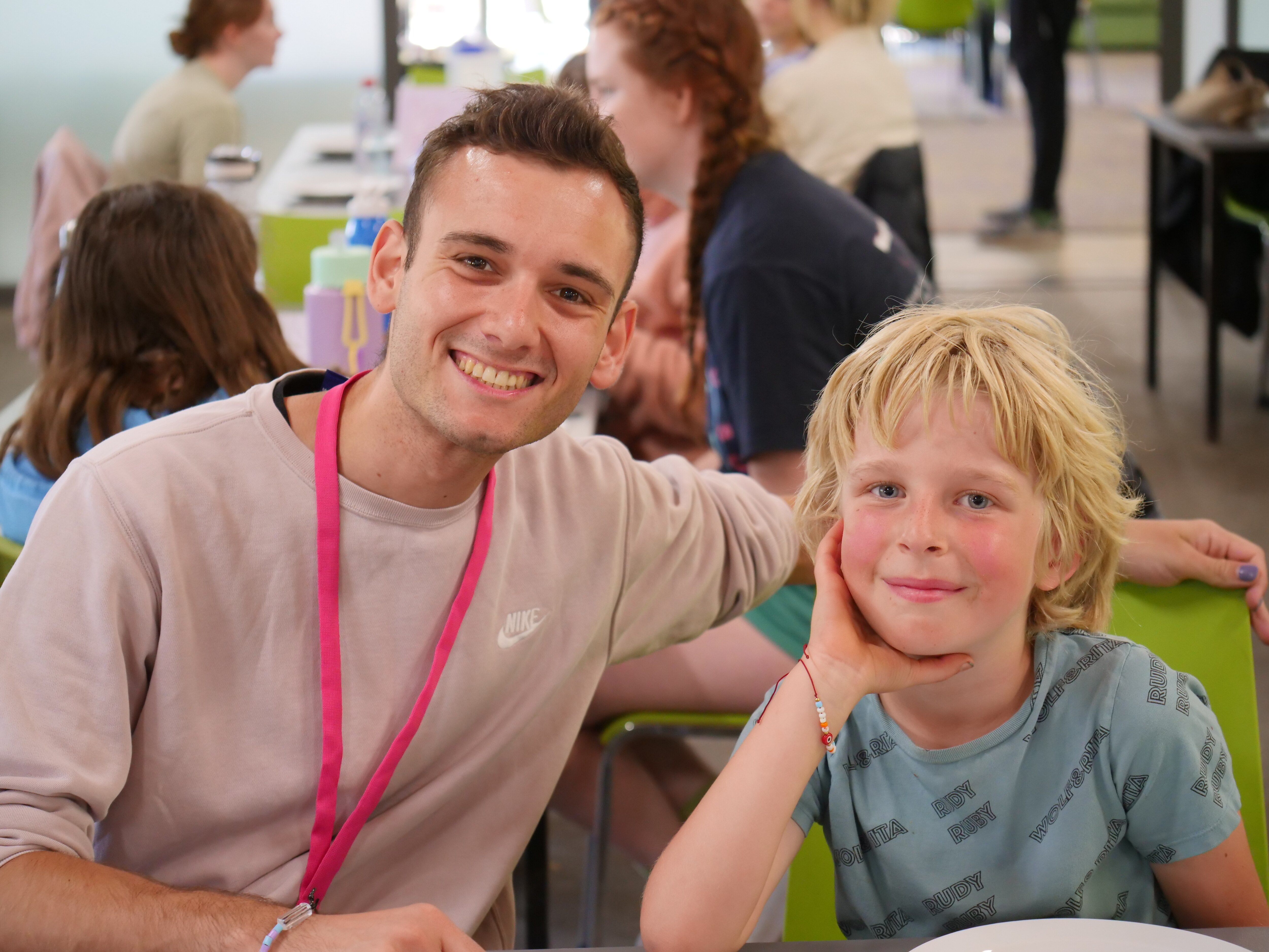 A man wearing a pink jumper and pink lanyard with short brown hair puts his arm behind a young boy with blond hair.