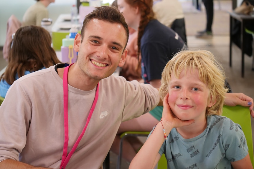 A man wearing a pink jumper and pink lanyard with short brown hair puts his arm behind a young boy with blond hair.