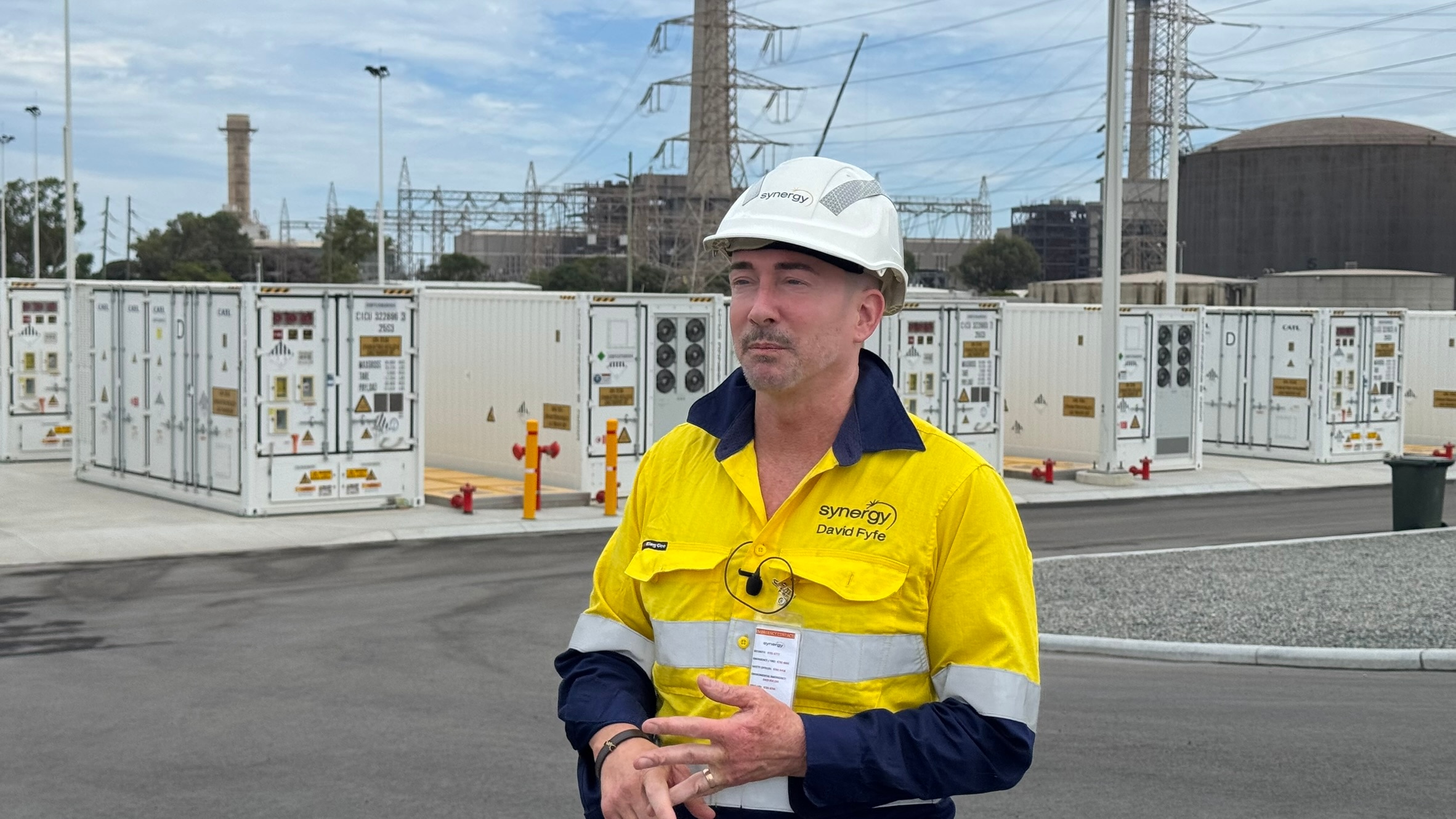 Man wearing yellow high-vis shirt and white hard-hat standing in front of batteries, power lines and old fuel tanks