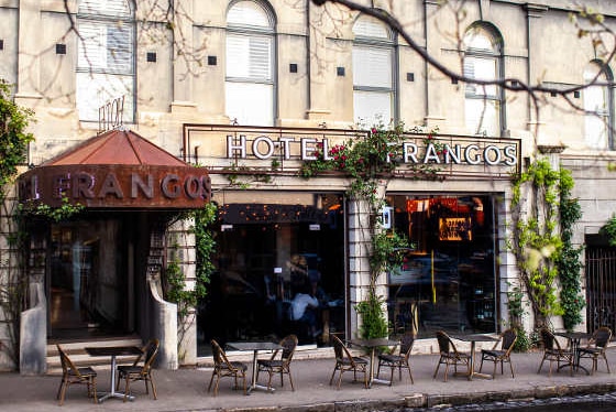 The exterior of a stone hotel with the words Hotel Frangos above the windows and seats and tables out the front.