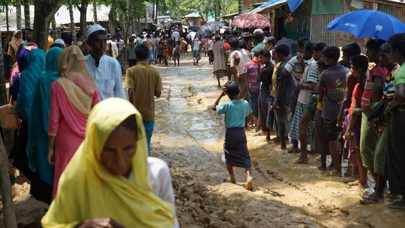 Refugees navigate a muddy path as others queue.