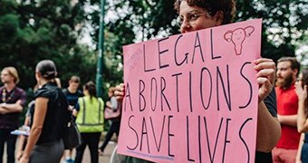 With a blurred crowd behind her, a woman holds a hand-written sign obscuring half her face, saying 'Legal abortions save lives'