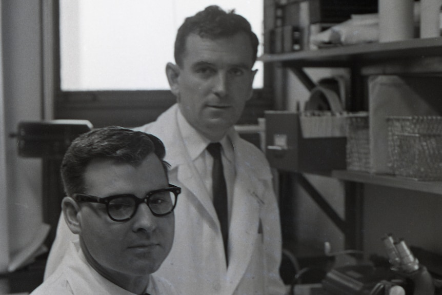 John Gorman and Vince Freda wearing white coats and ties in their lab in front of a microscope